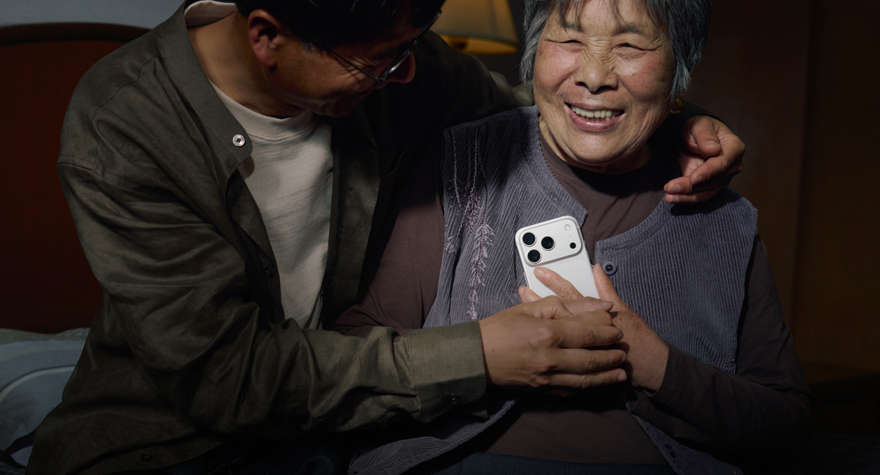 在房间里,中年儿子环抱着母亲,母亲双手将一部银色 iPhone 17 Pro 贴在胸前,脸上洋溢着开心的笑容。