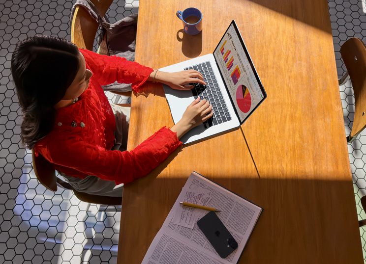 一个人坐在桌前使用 MacBook，一旁放着一个马克杯、一部智能手机和一些文件