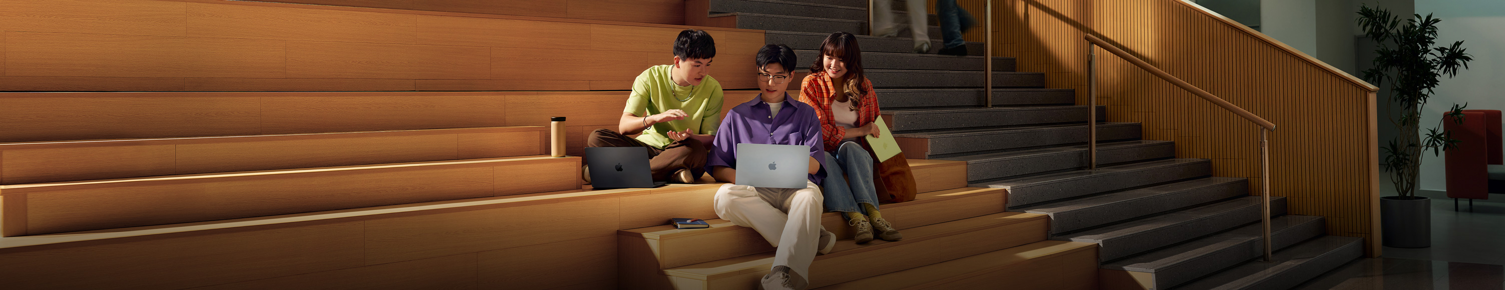三名学生坐在阶梯教室台阶上，一起看着中间那名学生手中打开的银色 MacBook Air 讨论。在他身边，一名学生拿着一台打开的深空黑色 MacBook Pro，另一名学生拿着合上的柑橘黄色 MacBook Neo。