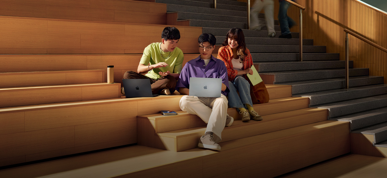 三名学生坐在阶梯教室台阶上，一起看着中间那名学生手中打开的银色 MacBook Air 讨论。在他身边，一名学生拿着一台打开的深空黑色 MacBook Pro，另一名学生拿着合上的柑橘黄色 MacBook Neo。