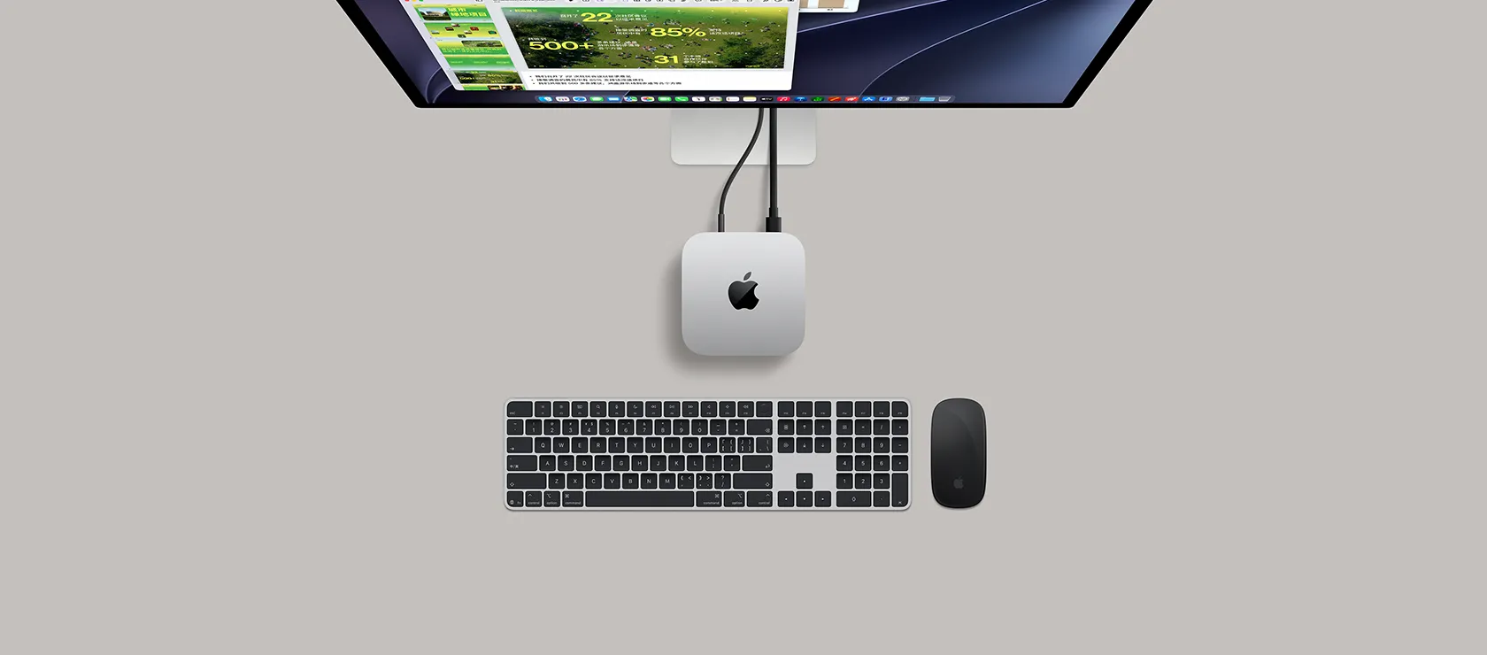Mac mini 的外观俯视图，展示银色铝金属机身、圆角设计，顶面居中有黑色亮面 Apple 标志，与线缆相连，上方的 Studio Display 部分显露出来，屏幕展示日历 app、Keynote 讲演演示文稿和条形图，下方是带有数字小键盘的妙控键盘和妙控鼠标。
