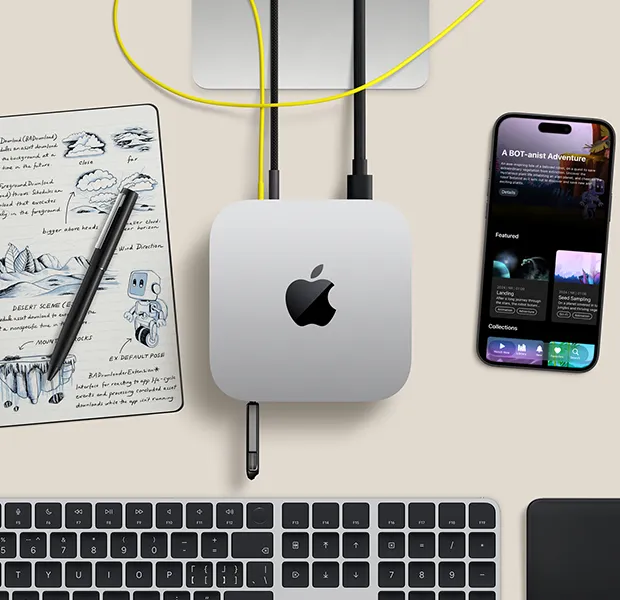 Mac mini 的外观俯视图，展示银色铝金属机身、圆角设计，顶面居中有黑色亮面 Apple 标志，与线缆相连，U 盘插入机身正面的 USB-C 端口，上方的 Studio Display 支架部分显露出来，周围环绕着 iPhone、妙控板、带有触控 ID 的妙控键盘、触控笔和电子墨水速绘板。