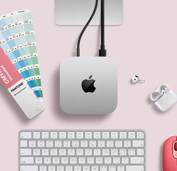 Mac mini 的外观俯视图，展示银色铝金属机身、圆角设计，顶面居中有黑色亮面 Apple 标志，与线缆相连，上方的 Studio Display 支架部分显露出来，周围环绕着 AirPods 4、Pantone 色卡，以及妙控键盘和红色 Logitech 鼠标。
