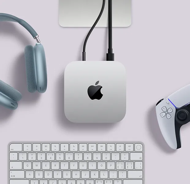 Mac mini 的外观俯视图，展示银色铝金属机身、圆角设计，顶面居中有黑色亮面 Apple 标志，与线缆相连，上方的 Studio Display 支架部分显露出来，周围环绕着蓝色 AirPods Max 2、白色 PlayStation DualSense 手柄和白色键盘。