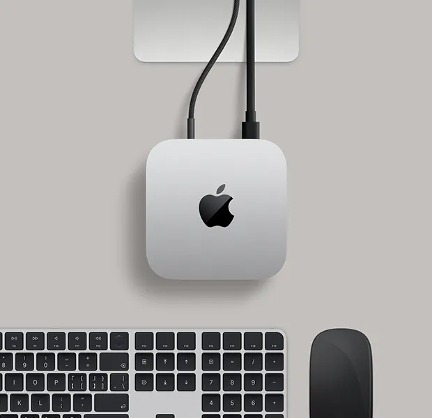 Mac mini 的外观俯视图，展示银色铝金属机身、圆角设计，顶面居中有黑色亮面 Apple 标志，上方的 Studio Display 支架部分显露出来，下方是带有触控 ID 的妙控键盘和妙控鼠标。