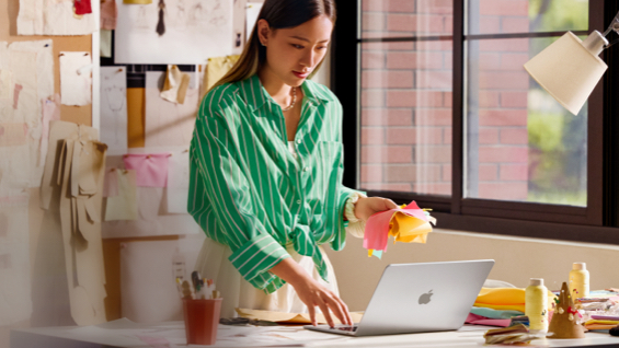 照片展示教室里，一位女生正站在书桌前使用 MacBook Air，背后的墙上贴满各种设计稿。