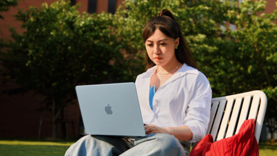 照片展示一位女生坐在草坪边的长椅上用着 MacBook Air。