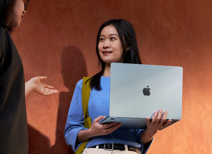 一名学生在户外与他人对话中，手里拿着打开的 MacBook Pro。
