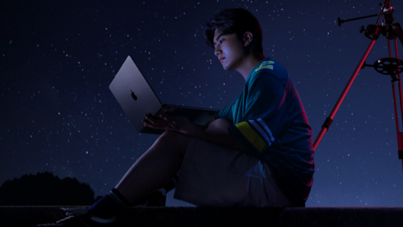 照片展示星空下，一位男生坐在天文望远镜前用着 MacBook Air。