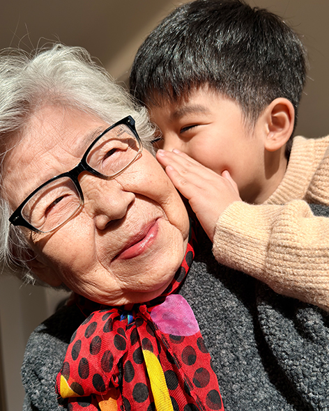 小朋友贴近外婆耳边轻声低语，祖孙二人沐浴在光线之中，脸上洋溢着幸福与温馨。由摄影师 randymu 用 iPhone 17 Pro 拍摄，使用珠光摄影风格。