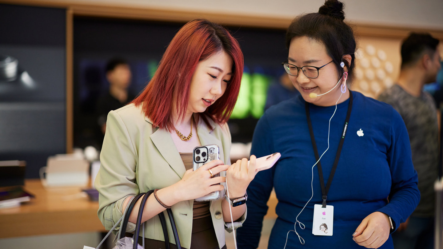 上海 Apple 南京东路零售店内，一位顾客手持粉色 iPhone 15 与团队成员交谈。