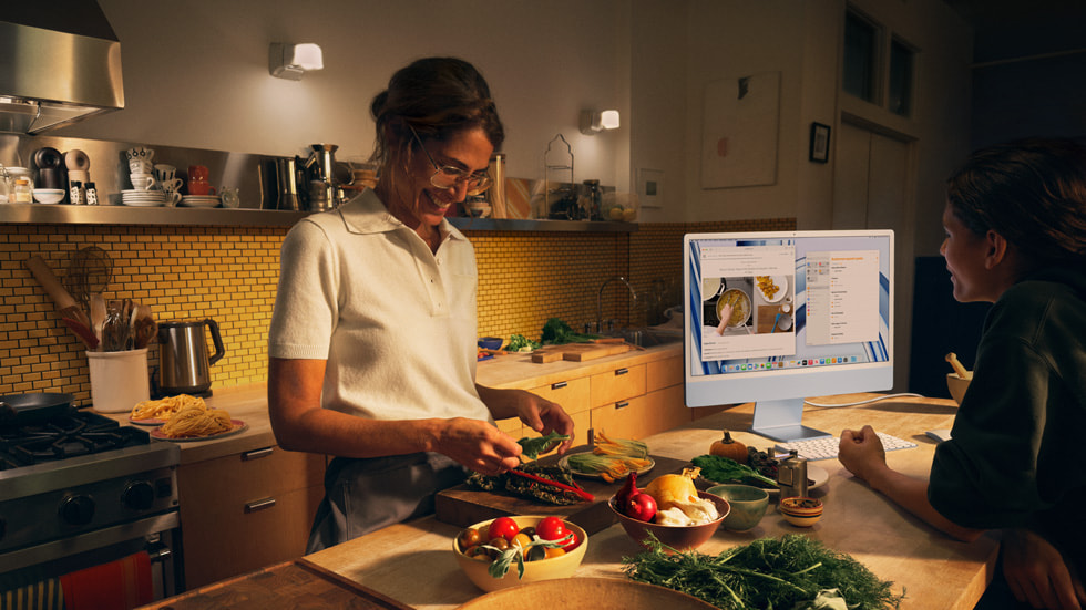 图中是一名家长和一名儿童，前者在厨房中烹饪，后者在使用搭载 M3 芯片的蓝色新款 iMac 以及同系列配色的键盘和鼠标。