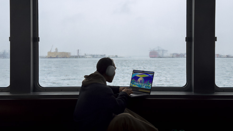 一位佩戴耳机、使用新款 MacBook Pro 的用户正望向窗外的港湾。