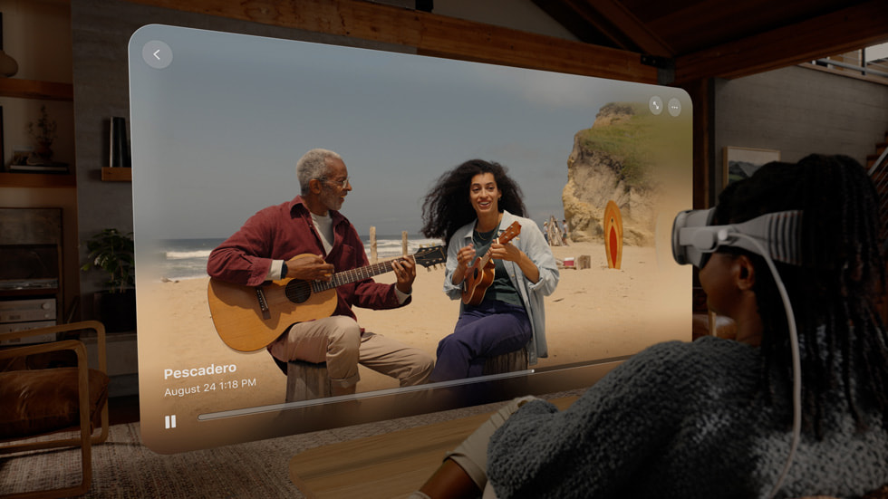 一个人佩戴 Apple Vision Pro，在客厅中观看视窗中放映的空间视频，视频中展现了两个人在海滩上播放音乐的场景。