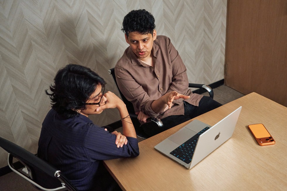 Shreela Agarwal 和导师 Chaitanya Tamhane 坐在桌旁，看着 MacBook Pro 进行讨论。iPhone 17 Pro Max 也放在一旁。