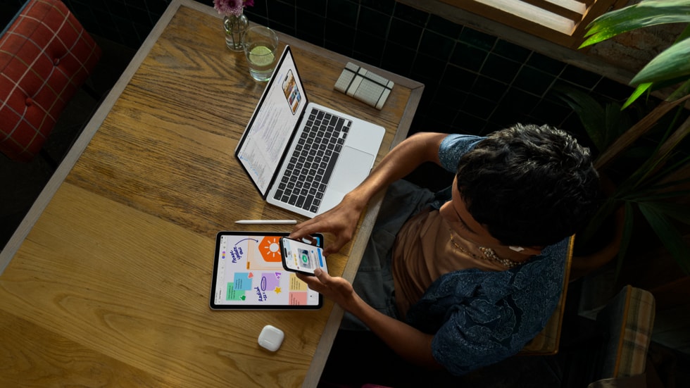 俯视图中是一个人手持 iPhone 在桌前使用 MacBook Pro 办公，设备屏幕上显示着编码界面，桌上还摆放着 iPad、Apple Pencil 和 AirPods。