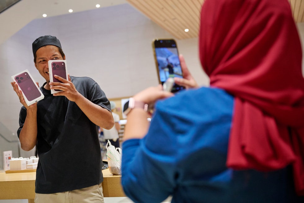 一位顾客在 Apple The Exchange TRX 零售店内拿着两台未拆封的 iPhone 18 Pro，另一个人正在用 iPhone 为前者拍照。
