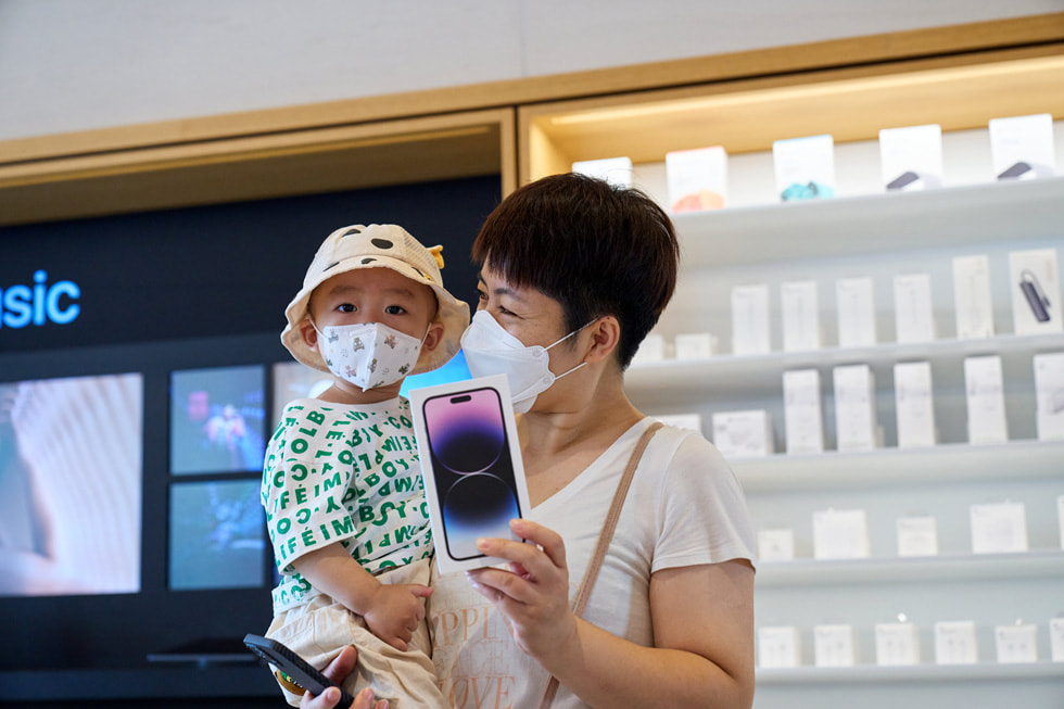 Apple 三里屯零售店内，一位顾客怀抱孩子，展示自己购买的 iPhone 14 Pro。