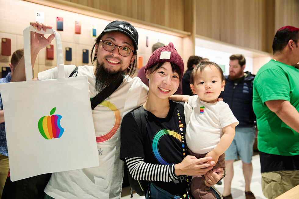 两位穿戴 Apple 设备的顾客举起一只 Apple 托特包，其中一位怀抱着婴儿。