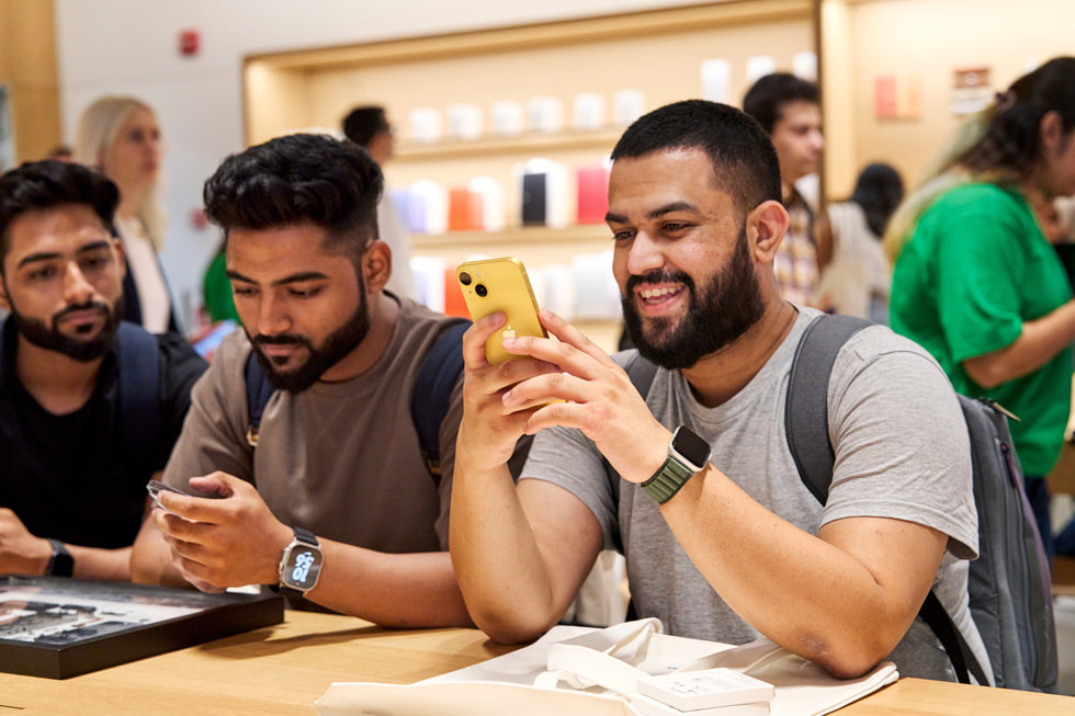 一位顾客坐在 Apple Saket 零售店内的展示桌旁，探索全新黄色 iPhone 14。