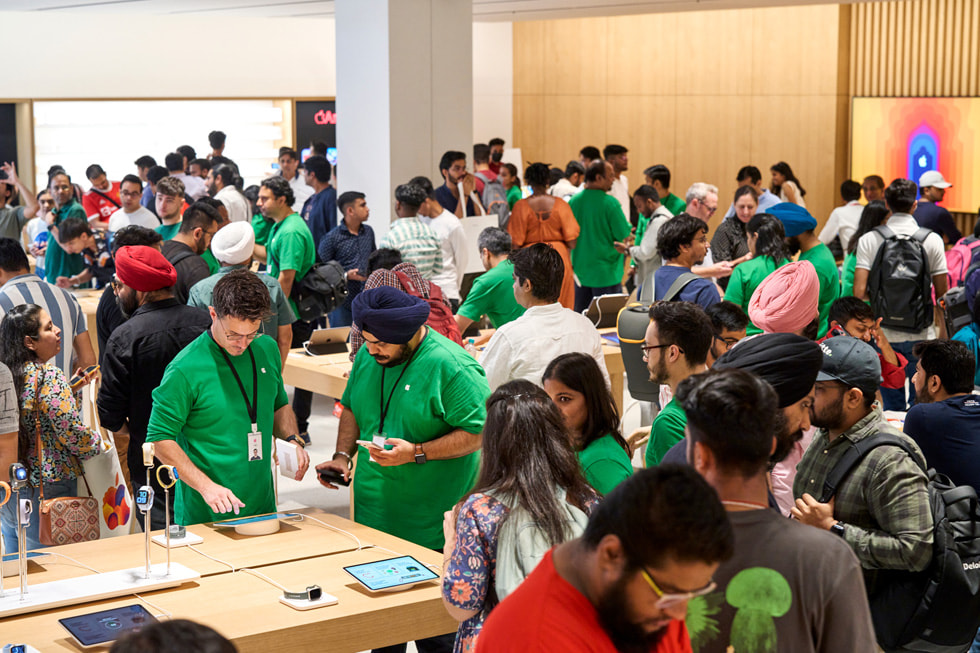 在 Apple Saket 零售店内，顾客与 Apple 团队成员交流，围绕在展示桌旁探索零售店的大量设备。