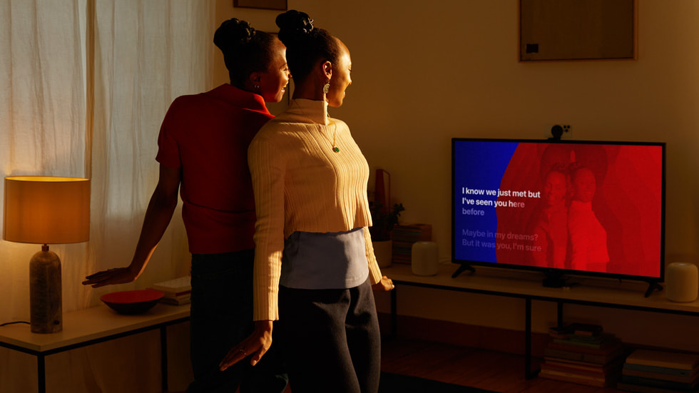 两位 Apple 用户背靠背站立，并同时出现在电视屏幕上。