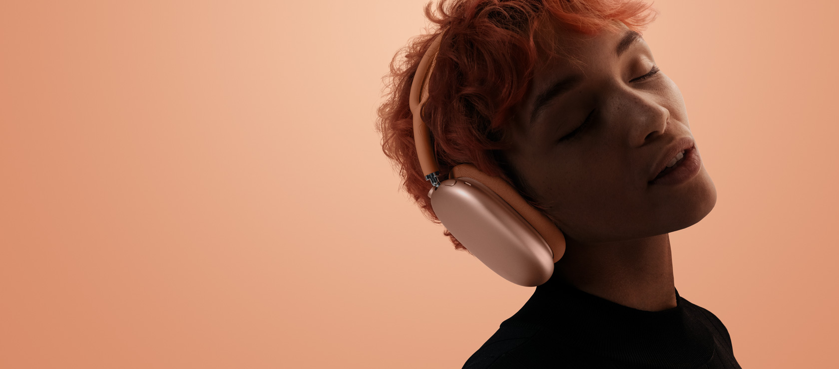 一个人戴着橙色 AirPods Max 2，闭着眼睛，头向一侧倾斜，其背景为橙粉色。