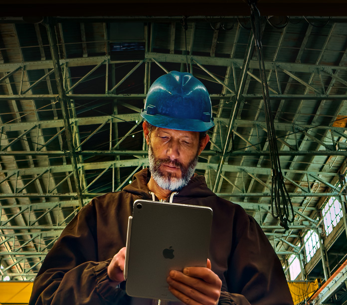 一个身穿棕色夹克、头戴蓝色安全帽的人，手中拿着吸附着 Apple Pencil 的银色 iPad，在头顶上方是金属横梁的工业环境中工作