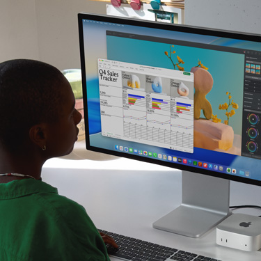 一名员工坐在 iMac 和 Mac Mini 前，使用它们处理一个名称为“第四季度销售追踪表”的 Excel 电子表格。这张 Excel 电子表格的后方是一个 Pixelmator Pro 窗口，以及 macOS Tahoe 背景。