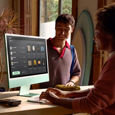 在一家售卖冲浪板和其他商品的店铺中，一位员工坐在柜台后面使用 iMac，为柜台另一侧的顾客结账。