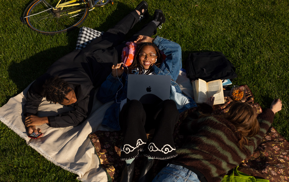 三名大学生躺在公园里的毯子上。一名学生手握 iPhone，一名学生在使用 MacBook Air 和 Apple 耳机，一名学生在翻看一本平装书。
