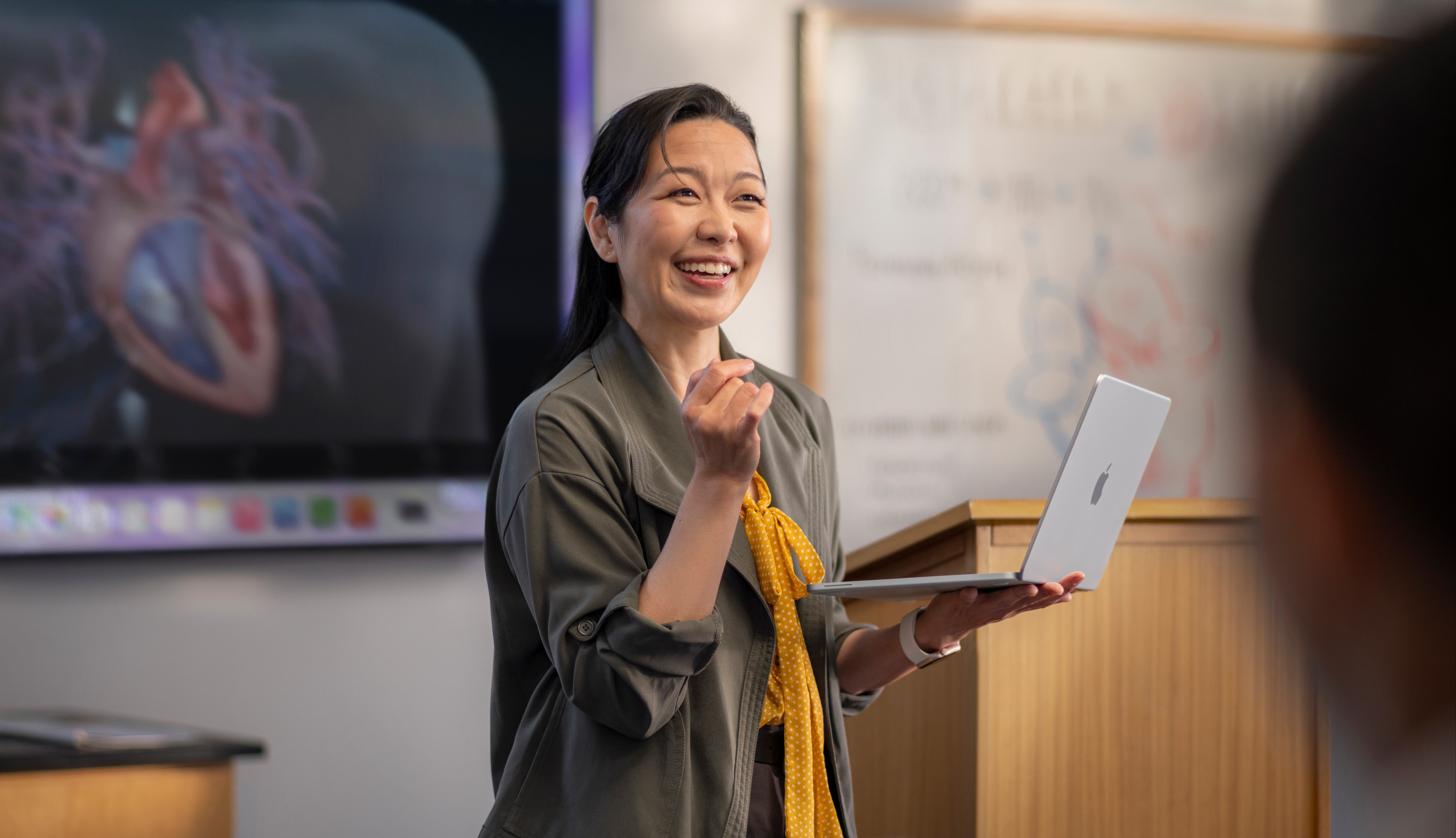 一位教授在报告厅做演讲，手上拿着 MacBook，展示性能和持久续航。
