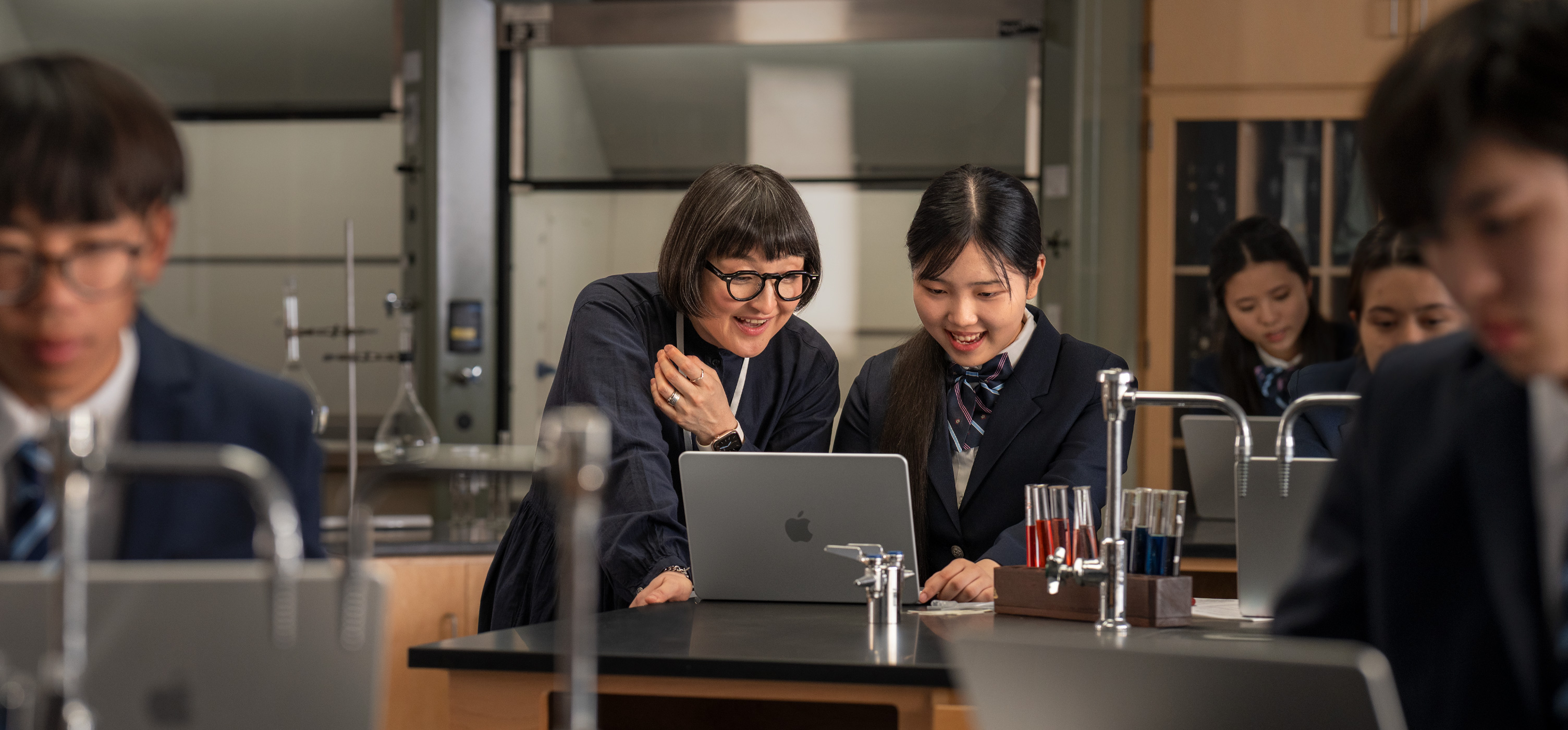 实验室环境中，学生和教师正在对着桌子上打开的 MacBook 进行互动，周围环绕着试管和设备。