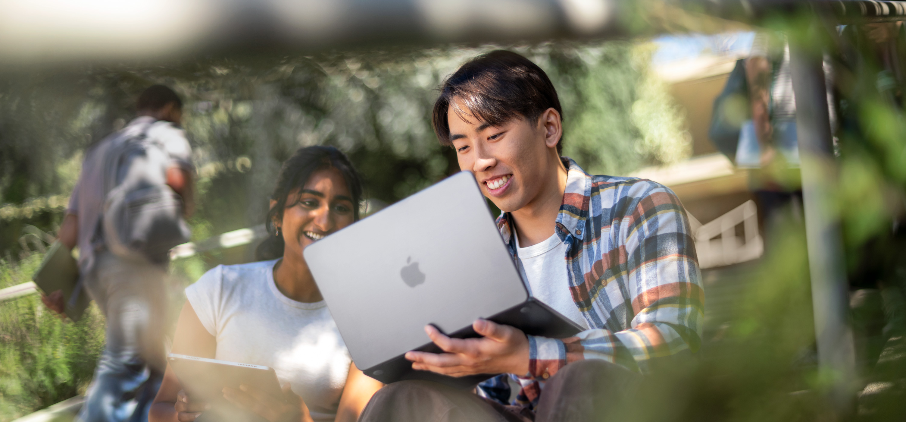 两位面带笑容的学生在户外使用 MacBook 协作完成作业，设备放在其中一人的腿上。