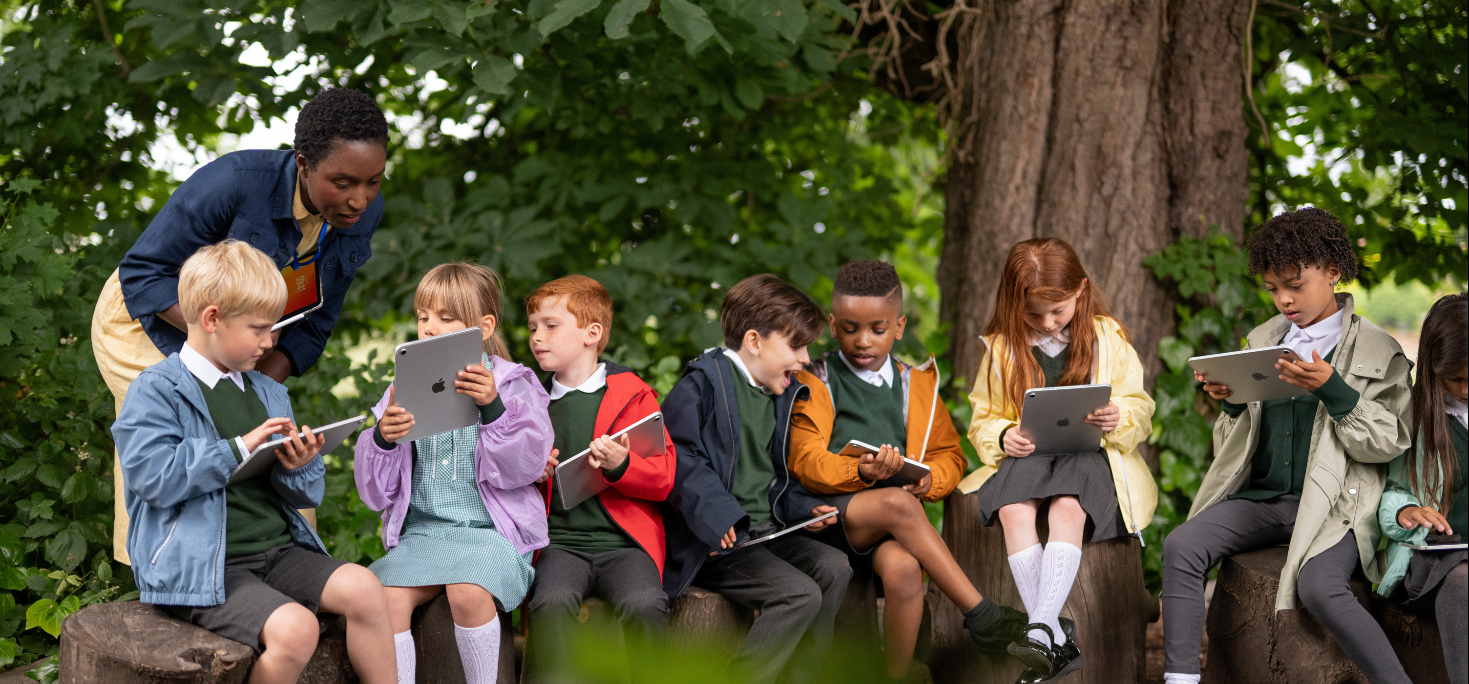 一群年幼的学生在户外使用 iPad，手拿 iPad 的教师则在指导他们，突显随时随地学习的灵活机动性。