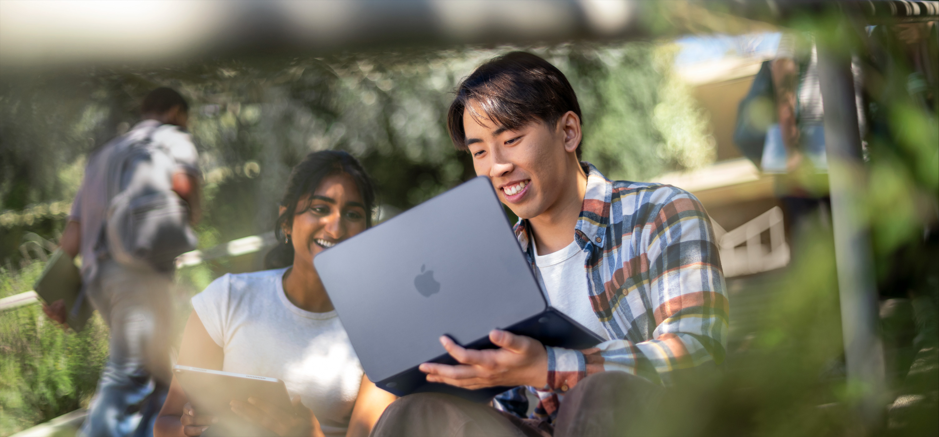 两位面带笑容的学生在户外使用 MacBook 协作完成作业，设备放在其中一人的腿上。
