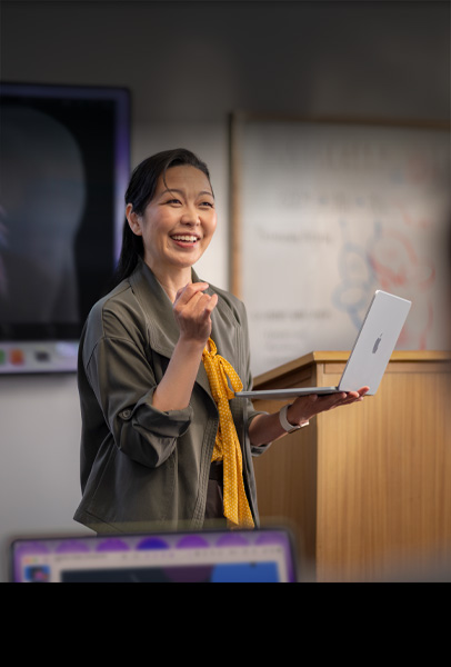 一位教授在报告厅为学生们做演讲，手上拿着银色的 MacBook，展示性能和持久续航力。