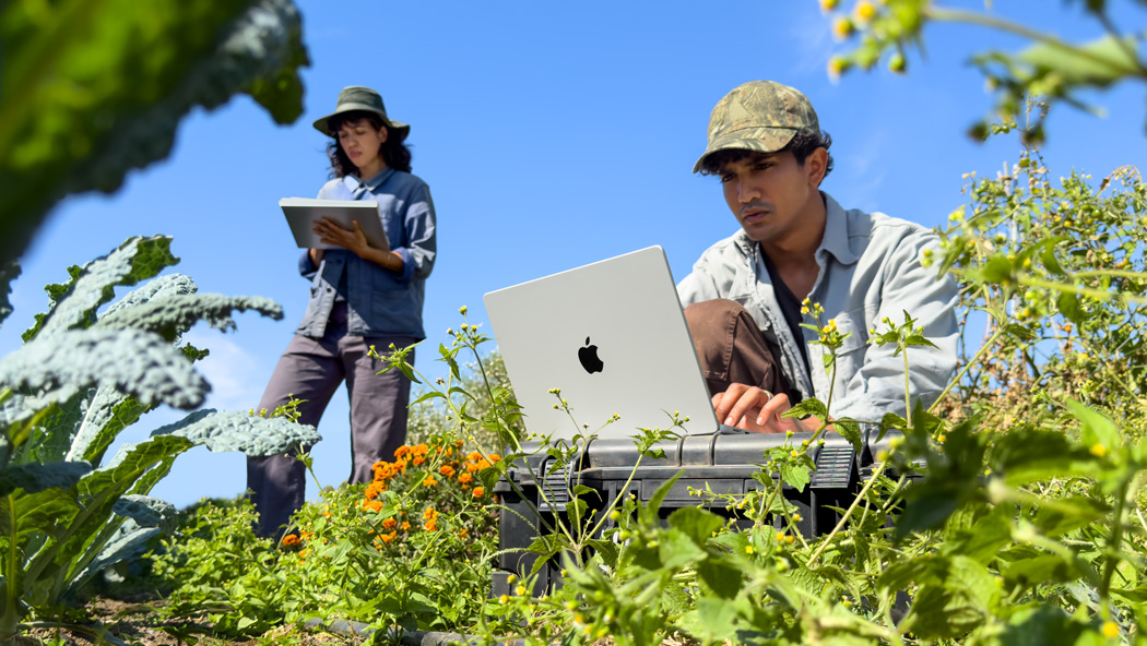 两个人在户外的花园里工作，其中一个人使用未接电源的银色 MacBook Pro，另一个人拿着记事本。